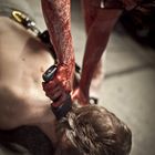 Blutverschmierte Arme halten einen Gegenstand über einem liegenden Mann mit heruntergelassenem Kopf.