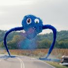 Ein großes, blaues, cartoonartiges Wesen mit langen Armen verfolgt eine Person auf einer ländlichen Straße.