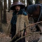 Ein Mann in traditioneller asiatischer Kleidung mit Hutmantel steht im Wald, ein Pferd mit Geschirr ist neben ihm.