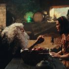 Eine alte Frau mit langen weißen Haaren reicht einem jungen Mädchen einen roten Apfel in einer rustikalen, gemütlichen Hütte.