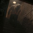 Ein Mann trägt eine Person auf dem Rücken eine Treppe hinauf, umgeben von Ziegelwänden, schwach beleuchtet.