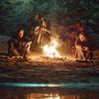 Zwei Personen sitzen am Lagerfeuer im Wald, mit Motorrädern im Hintergrund. Sandiger Boden reflektiert das Feuer.