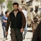 Ein Mann mit Bart geht entschlossen eine staubige Straße entlang, während Menschen im Hintergrund zu sehen sind.