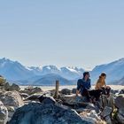 Zwei Personen sitzen auf Felsen in einer Berglandschaft, umgeben von Schnee und blauem Himmel.