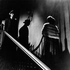 Ein Mann im Anzug und eine Frau im Kleid stehen auf einer Treppe, beleuchtet von dramatischem Licht und Schatten.