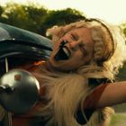 Person mit blonden Haaren lehnt aus einem Auto, Mund weit offen, wirkt fröhlich oder ekstatisch. Hintergrund bewaldet.