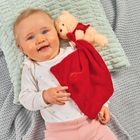 Kein Text. Ein fröhliches Baby liegt auf einer Decke, hält einen roten Stoff und ein Kuscheltier.