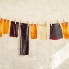 Stoffstücke in dunklen und warmen Tönen hängen an einer Leine mit Klammern. Heller, texturierter Hintergrund.