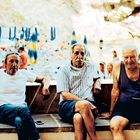 Drei ältere Männer sitzen auf einer Mauer. Im Hintergrund sieht man einen Strand mit blauen Sonnenschirmen.