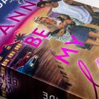 Text: "Wanna BE MY lover" in großen pinken Buchstaben. Illustration eines jungen Paares vor einem Haus mit Palmen und Autos.