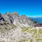 Keine Texte zu erkennen. Eine Berghütte vor dramatischer Gebirgskulisse mit blauem Himmel und Wanderpfaden auf steinigem Gelände.