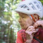 Eine ältere Person trägt einen weißen Helm und bindet den Kinnriemen fest. Sie befindet sich im Freien mit Bäumen im Hintergrund.