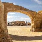 Lehmstrukturen bilden eine bogenförmige Szenerie in einer Wüstenlandschaft unter einem klaren blauen Himmel.