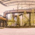 Die Szene zeigt ein Industriegelände mit alten Fabrikgebäuden, Rohren und jungen Bäumen.