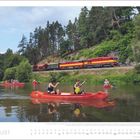 AUGUST. Menschen paddeln in roten Kanus auf einem Fluss; im Hintergrund fährt ein rot-gelber Zug vorbei.