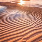 Kalender für August, zeigt einen Sonnenuntergang am Strand mit wellenförmigen Sandstrukturen und warmen Farben.
