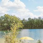Kein sichtbarer Text. See mit grünem Ufer, Bäumen und gelbem Sonnenschirm am Wasser, blauer Himmel mit weißen Wolken.