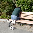 Eine Person kauert auf einer Parkbank, Schuhe am Boden, umgeben von dichtem Grün.
