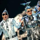 Drei Personen in silberglänzender Kleidung. Einer trägt eine Uniform mit "18". Alle tragen Sonnenbrillen und extravagante Hüte.