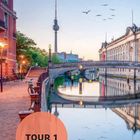 Text "TOUR 1" in einem orangefarbenen Kreis. Fluss mit Brücke, Fernsehturm, Gebäude und fliegende Vögel im Hintergrund.