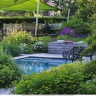 Kalenderwoche 26, naturreiche Terrasse mit Holztisch und Stühlen, umgeben von üppigem, grünem Garten im Sonnenlicht.