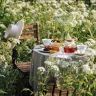 Keine sichtbaren Texte. Ein Picknicktisch in einem blühenden Feld, gedeckt mit Tee, Kuchen und Blumen, daneben ein Holzstuhl.