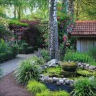 Ein ruhiger Garten mit einem kleinen Springbrunnen, umgeben von Steinen, Blumen und Bäumen, neben einem Häuschen mit Ziegeldach.
