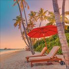 Zwei Liegestühle mit rotem Sonnenschirm am Strand, umgeben von Palmen bei Sonnenuntergang. Links ein Steg ins Meer.