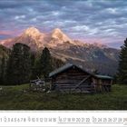Kalenderseite für August 2023: Eine Holzhütte vor einem Bergmassiv bei Sonnenuntergang, umgeben von Wald.
