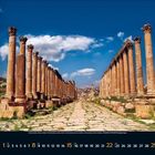 Augustkalender mit antiken Säulenreihen auf altem gepflastertem Weg, blauer Himmel mit Wolken, grüne Landschaft.