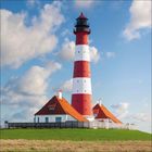 Ein rot-weiß gestreifter Leuchtturm steht auf einem grünen Hügel, umgeben von Gebäuden mit roten Dächern unter blauem Himmel.