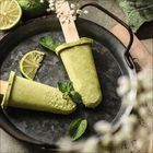 Zwei grüne Eis am Stiel auf einer Metallplatte mit Limettenscheiben und Minzblättern, in einer rustikalen Umgebung.