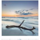 Kalender August mit Foto: Ein sonnenuntergang am Meer, ein hölzernes Treibholz liegt im Sand vor sanften Wellen.