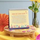 Auf einem Tisch steht ein Kalender mit dem Text "NIMM DAS LEBEN LEICHT". Bunte Blumen aus Papier sind dekorativ verteilt.