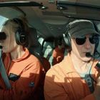 Zwei Personen in orangefarbener Kleidung sitzen im Cockpit eines Flugzeugs, halten Ferngläser und tragen Headsets.