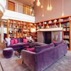 Ein gemütlicher Raum mit violetten Sofas, Kissen, hölzernen Bücherregalen, Kamin und hängenden Glühbirnen.