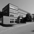 Ein modernes, mehrstöckiges Backsteingebäude mit vielen Fenstern, umgeben von Bäumen und einer leeren Straße.