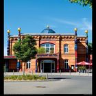 "BAHNHOF UELZEN" steht auf dem Eingang eines architektonisch auffälligen Gebäudes mit bunten Säulen. Darunter stehen Zahlen von 1 bis 31; VIII 2027.