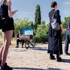 Text: Adrien Missika, MOTUS, pop-up exhibition, feat. Geoffrey Hendricks in front of Berghain, 17.7.2022.  
Mehrere Menschen stehen draußen; ein Mann sitzt mit einem blauen Kunstwerk auf einem Fahrradanhänger.