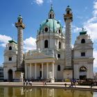 Kirche mit grünem Kuppeldach, flankiert von zwei verzierten Säulen. Menschen spazieren an einem Wasserbecken entlang.