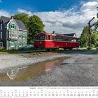 Wuppertal – Triebwagen VT 95. August 2027 Kalender. Ein roter Zug steht vor einem alten, grauen Haus neben grünen Bäumen.
