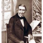 Ein älterer Herr sitzt vor einem Tisch mit Zeichnungen von Vögeln; links ein Fenster mit Pflanzen.