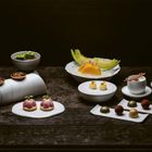 Verschiedene kunstvolle Dessert-Kreationen auf weißen Tellern. Schalen mit Pralinen, Melonenscheiben, kleine Törtchen, dekorative Details.