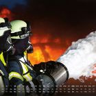 Kalendertage und Wochentage unten, zwei Feuerwehrleute in Schutzkleidung löschen ein Feuer mit Schaum.