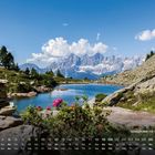 "Spiegelsee mit Dachstein 9." Ein Bergsee, umgeben von Felsen, Bäumen und bunten Blumen, mit hohen Bergen im Hintergrund.