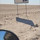 Text "ICE COLD BEER!!!" auf einem Schild in einer sandigen Wüstenlandschaft. Ein Rückspiegel zeigt einen Teil eines Fahrzeugs.