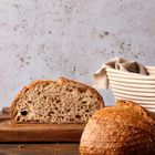 Ein Laib Sauerteigbrot, angeschnitten, liegt auf einem Holzbrett. Daneben ein Messer und ein leerer Brotkorb.