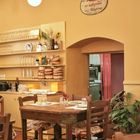 Griechischer Text auf einem ovalen Schild an gelber Wand, darunter ein rustikales Restaurantinterieur mit Holztischen.
