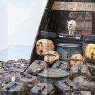 Text "50" auf Objekten; skulpturale Anordnung von Kanistern und Fässern, mit einer Figur oben drauf.
