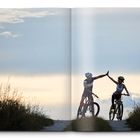 Zwei Radfahrer auf einem Feldweg geben sich High-Five, umgeben von Gras und einem bewölkten Himmel.